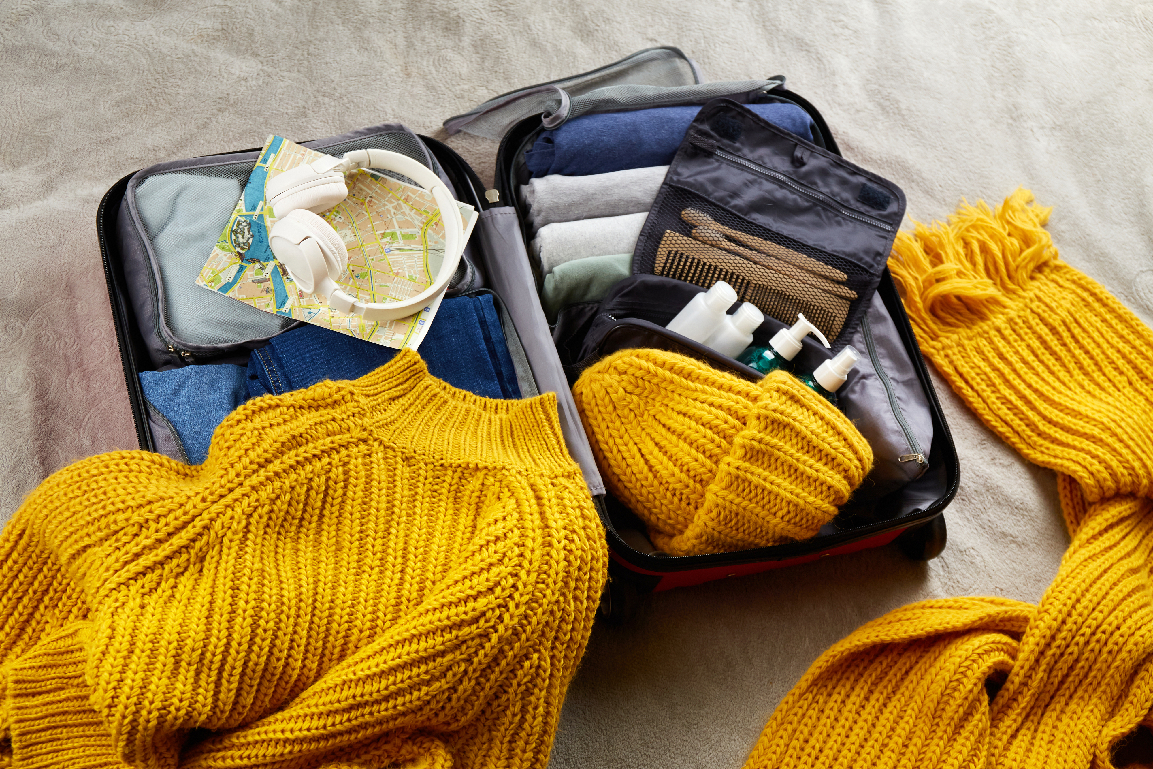 Open suitcase with clothes, headphones, map, and toiletries, alongside a yellow knitted scarf and hat
