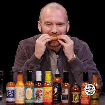 Man eating a wing at a table with hot sauce bottles, logo &#x27;First We Feast&#x27; visible