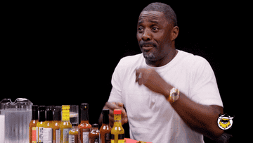 Man in a white shirt gesturing with a surprised expression sitting at a table with hot sauce bottles