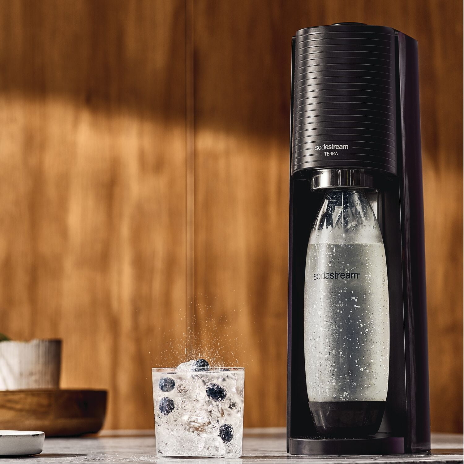 SodaStream machine carbonating water in a bottle next to a glass with fizzing water and ice cubes, placed on a wooden surface