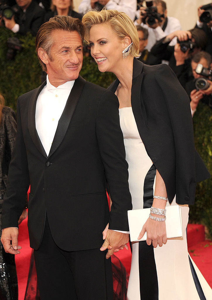 Both holding hands on the red carpet, with Sean in classic tuxedo and Charlize in elegant dress with cape jacket; photographers in background