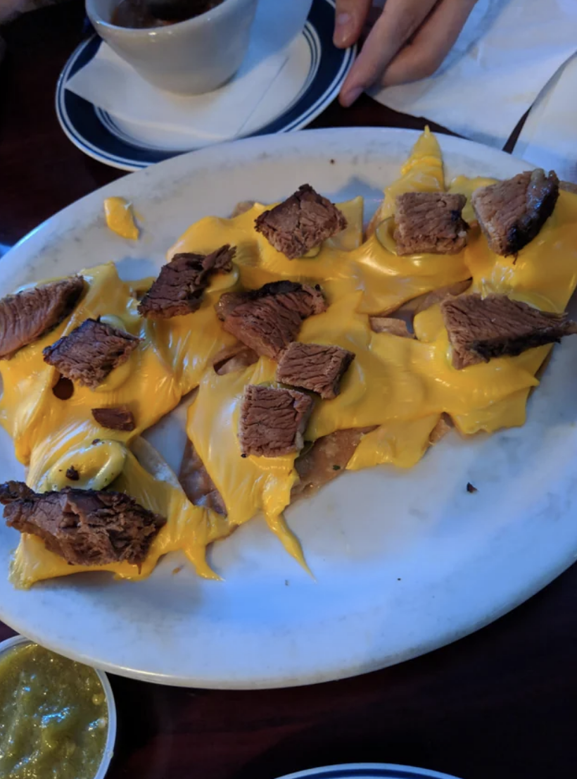 A plate of nachos with partially melted American cheese and dry beef toppings, served on a table with a side of green salsa