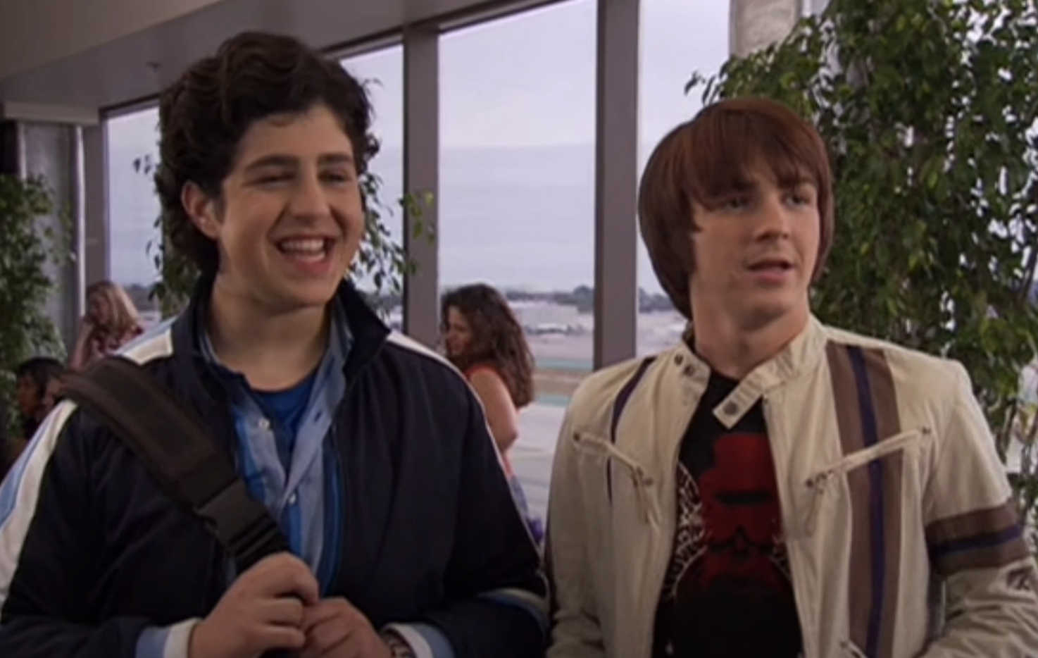 Two characters from a TV show, one with a backpack and the other in a jacket, are indoors with a view of the ocean through a window behind them