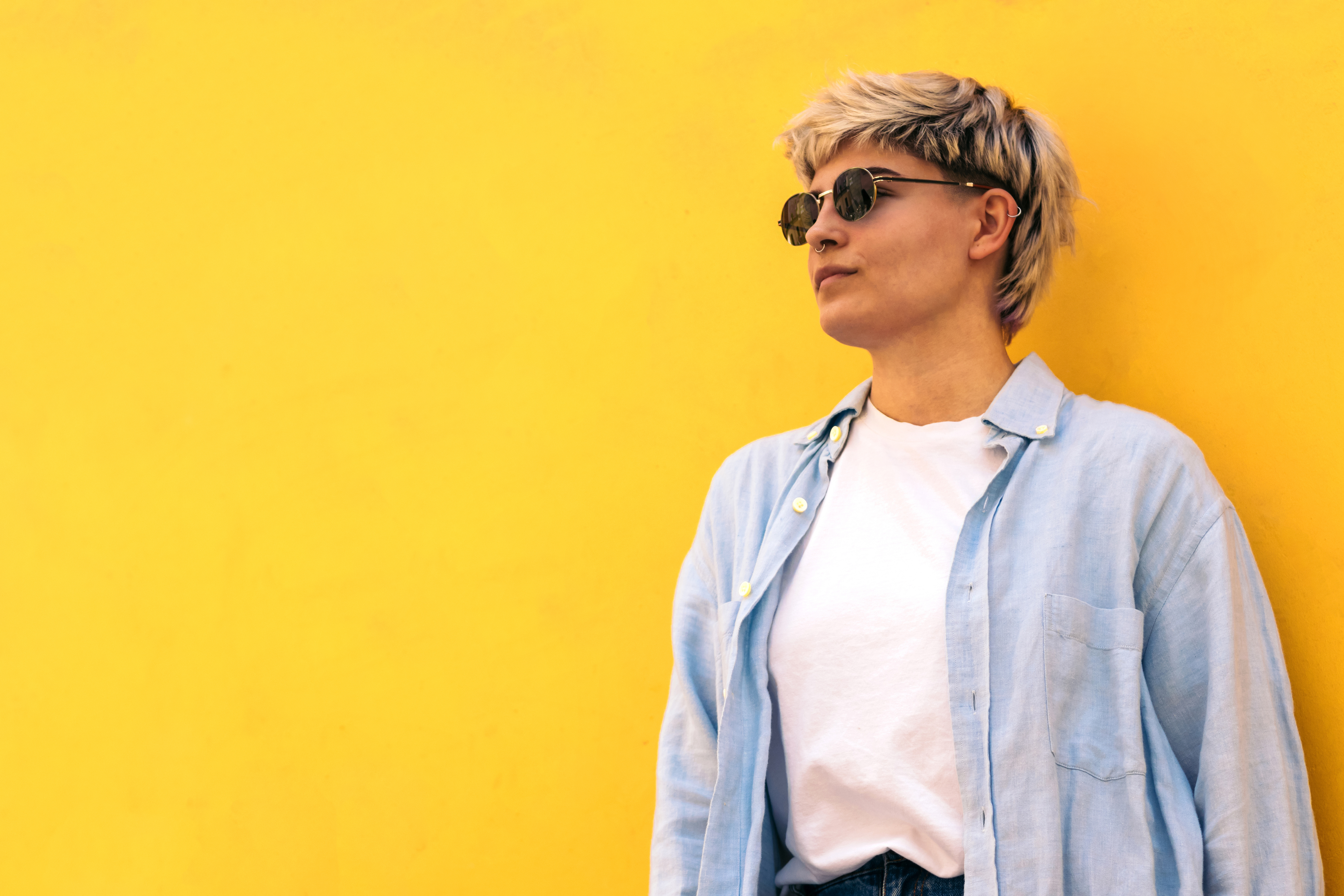 Woman wearing sunglasses and casual shirt standing by a yellow wall, looking to the side