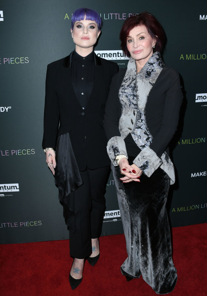 Kelly and Sharon Osbourne standing side by side; Kelly sports a black outfit with a unique haircut, Sharon in a textured jacket and pants