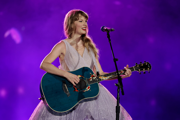 Taylor Swift singing and playing guitar on stage with a purple backdrop. She wears a glittery dress