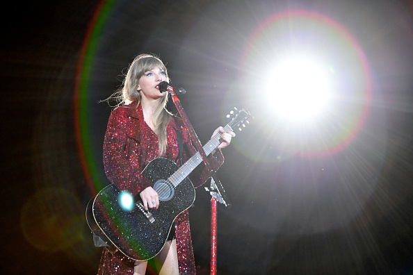 Taylor Swift performs with guitar, wearing a glittery outfit with bright stage lights behind her