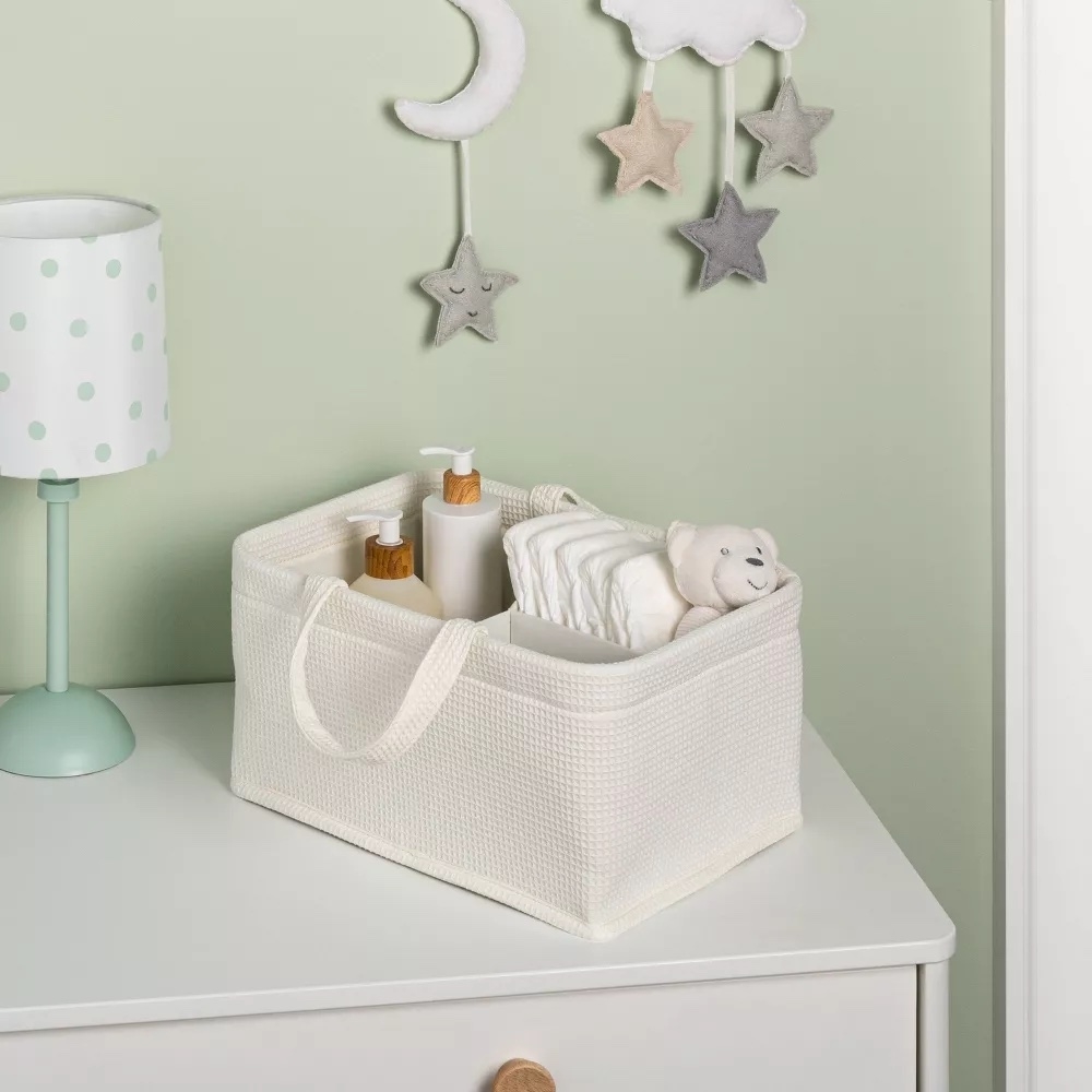 Woven diaper caddy with baby items on a dresser next to a lamp