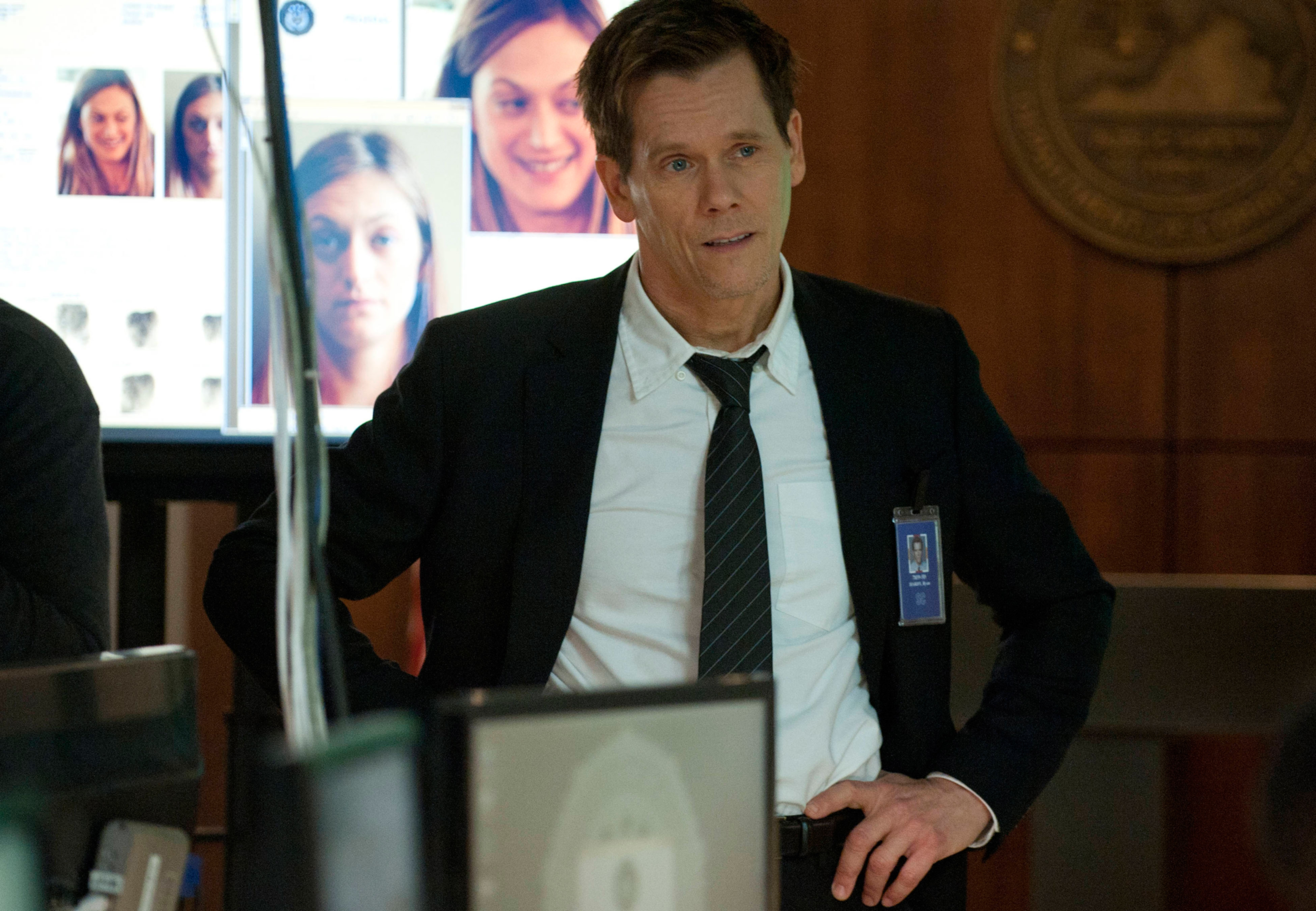Kevin Bacon in a suit with badge, portraying an FBI agent in a film, with monitors displaying faces in the background