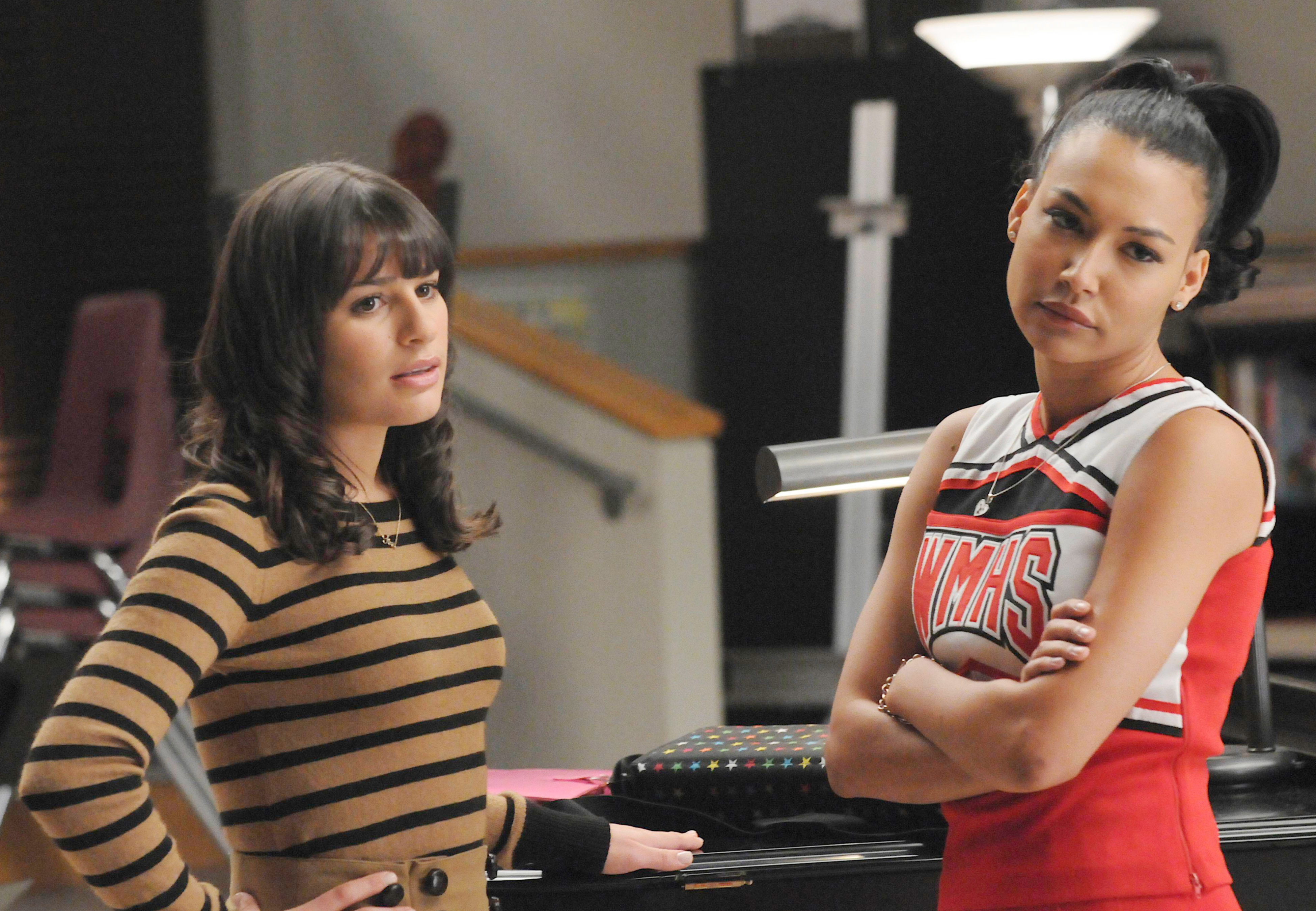 Two characters from Glee, Rachel in a striped top and Santana in a cheerleader outfit, stand facing each other with serious expressions