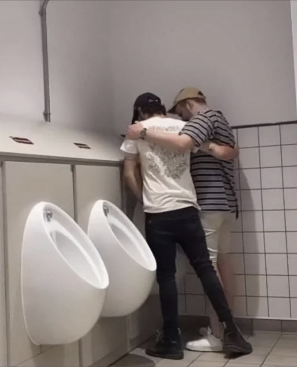 Two men jokingly pose using the same urinal