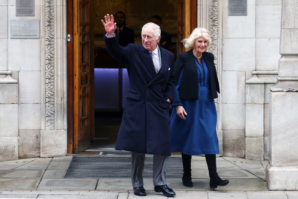 Prince Charles and Camilla exiting a building, he waves in a coat, she smiles beside him