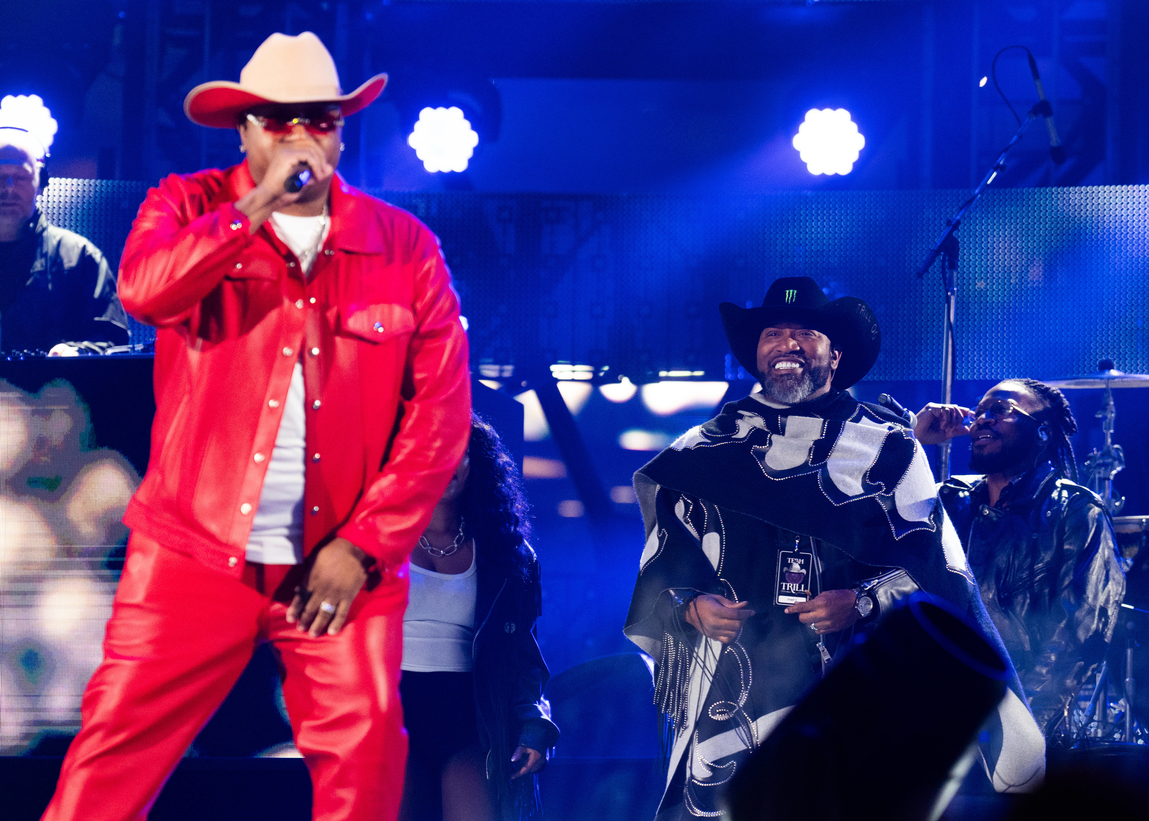 Two musicians in cowboy hats perform on stage; one in a red outfit with a microphone, the other in a black and white poncho