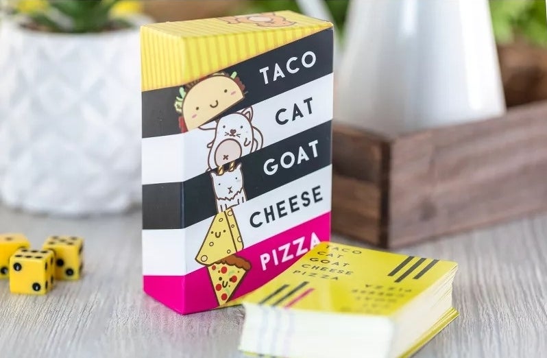 A card game named &quot;TACO CAT GOAT CHEESE PIZZA&quot; with whimsical illustrations, set on a table next to dice