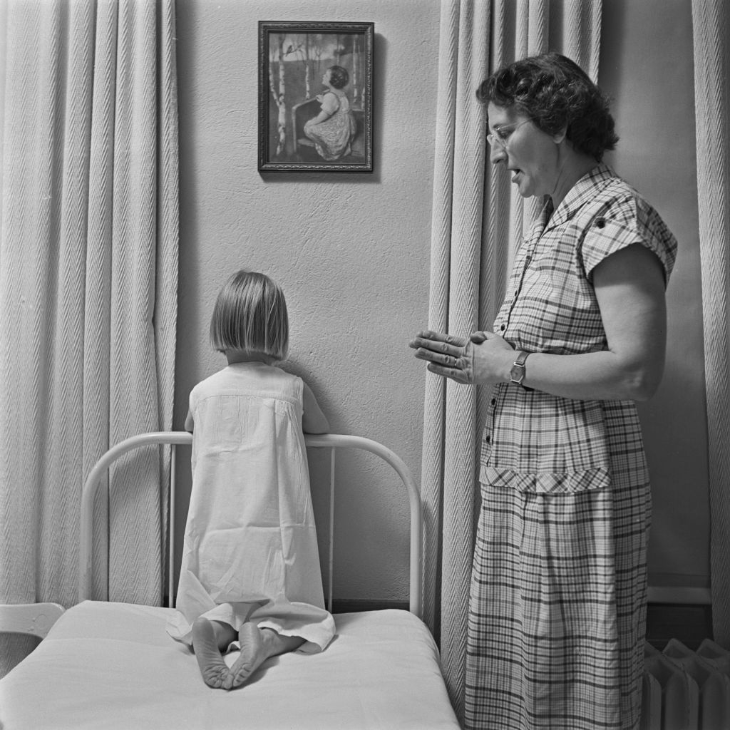 Woman standing by a child sitting on a bed, facing a wall, both in vintage attire