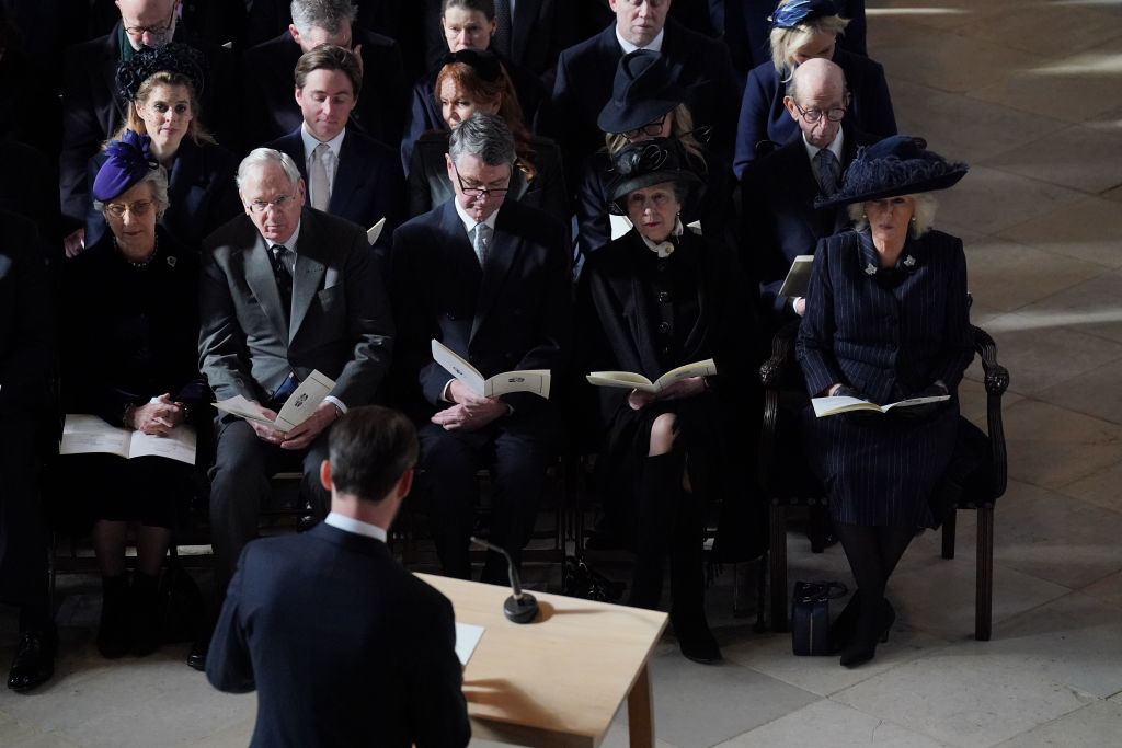 A group of people, including royalty, seated at a funeral, listening to a speaker at a podium
