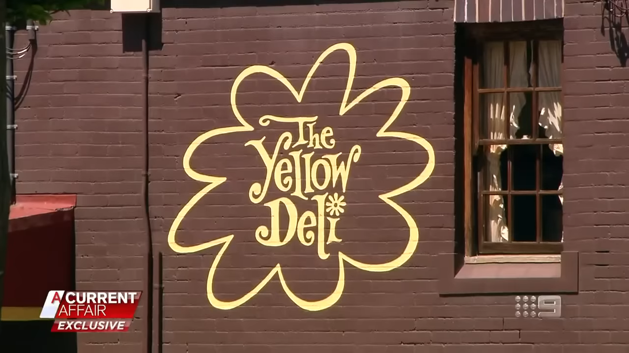 A sign on a building reads &quot;The Yellow Deli&quot; with a decorative flower around it