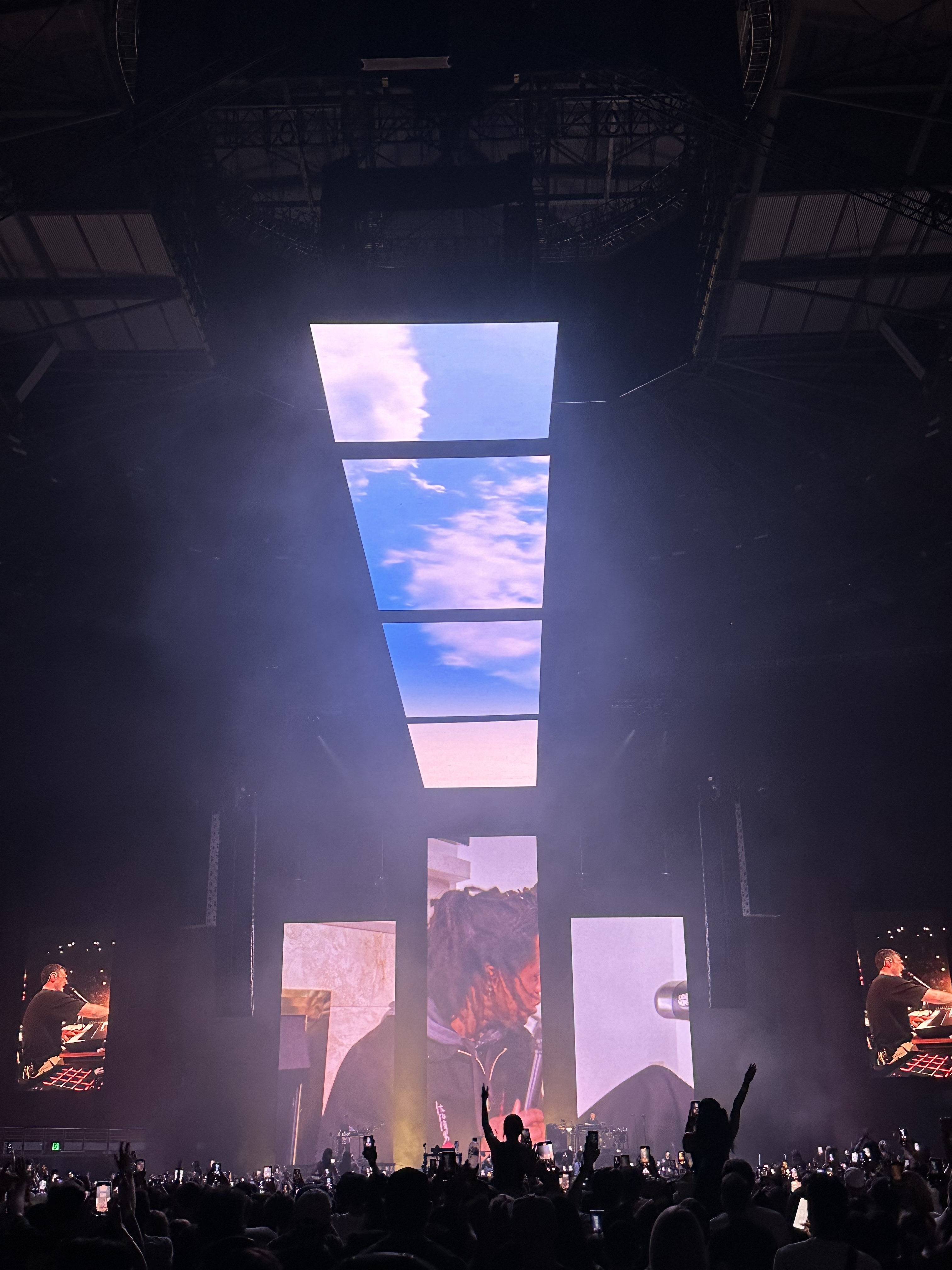 Concert stage with large screens showing sky and nature, audience with hands up in silhouette