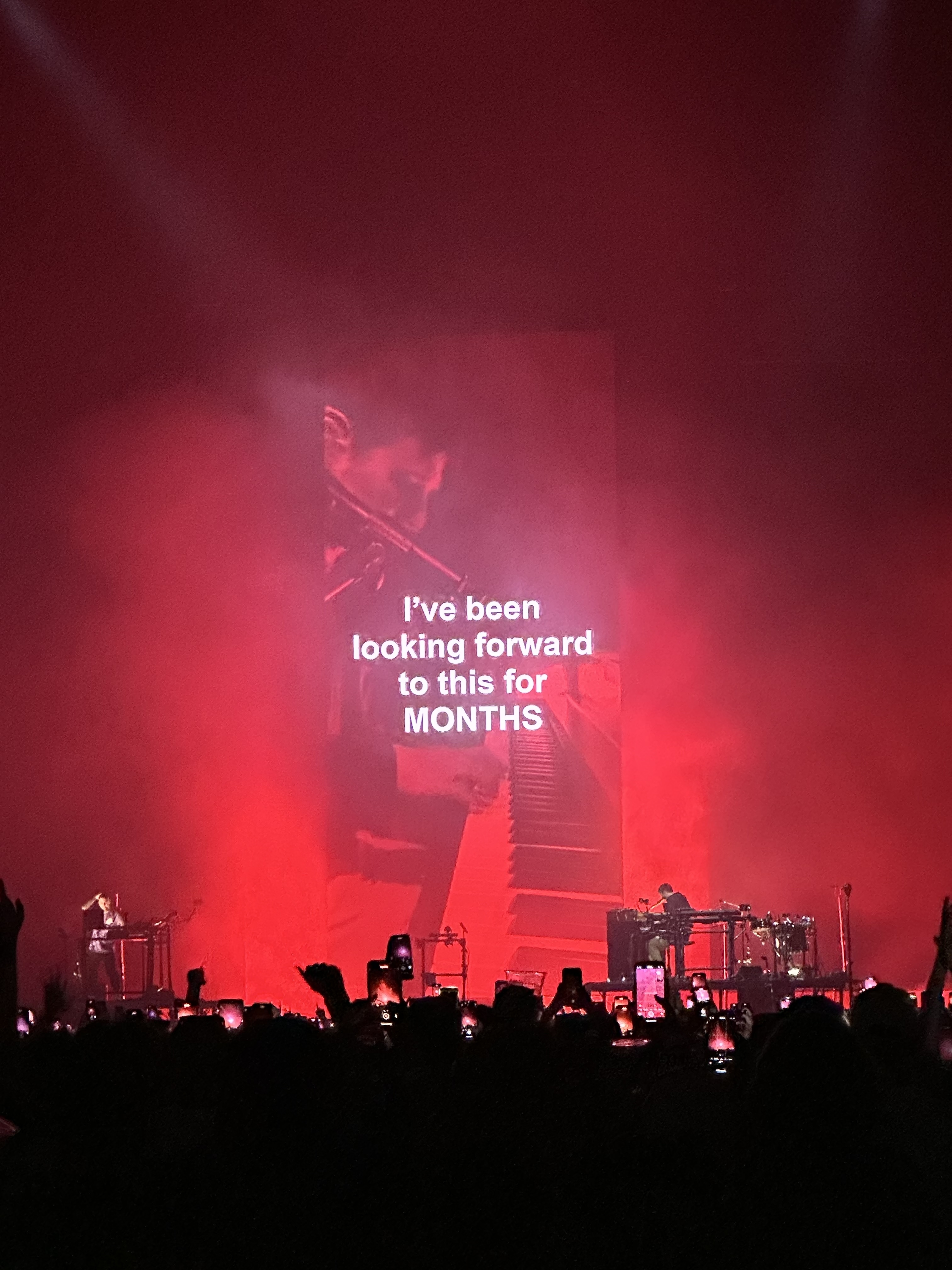 Concert stage with a large screen displaying a message, &quot;I&#x27;ve been looking forward to this for MONTHS,&quot; silhouettes of crowd and band