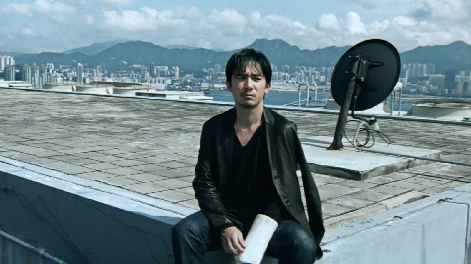 Man in black jacket sitting on rooftop with satellite dish and cityscape behind