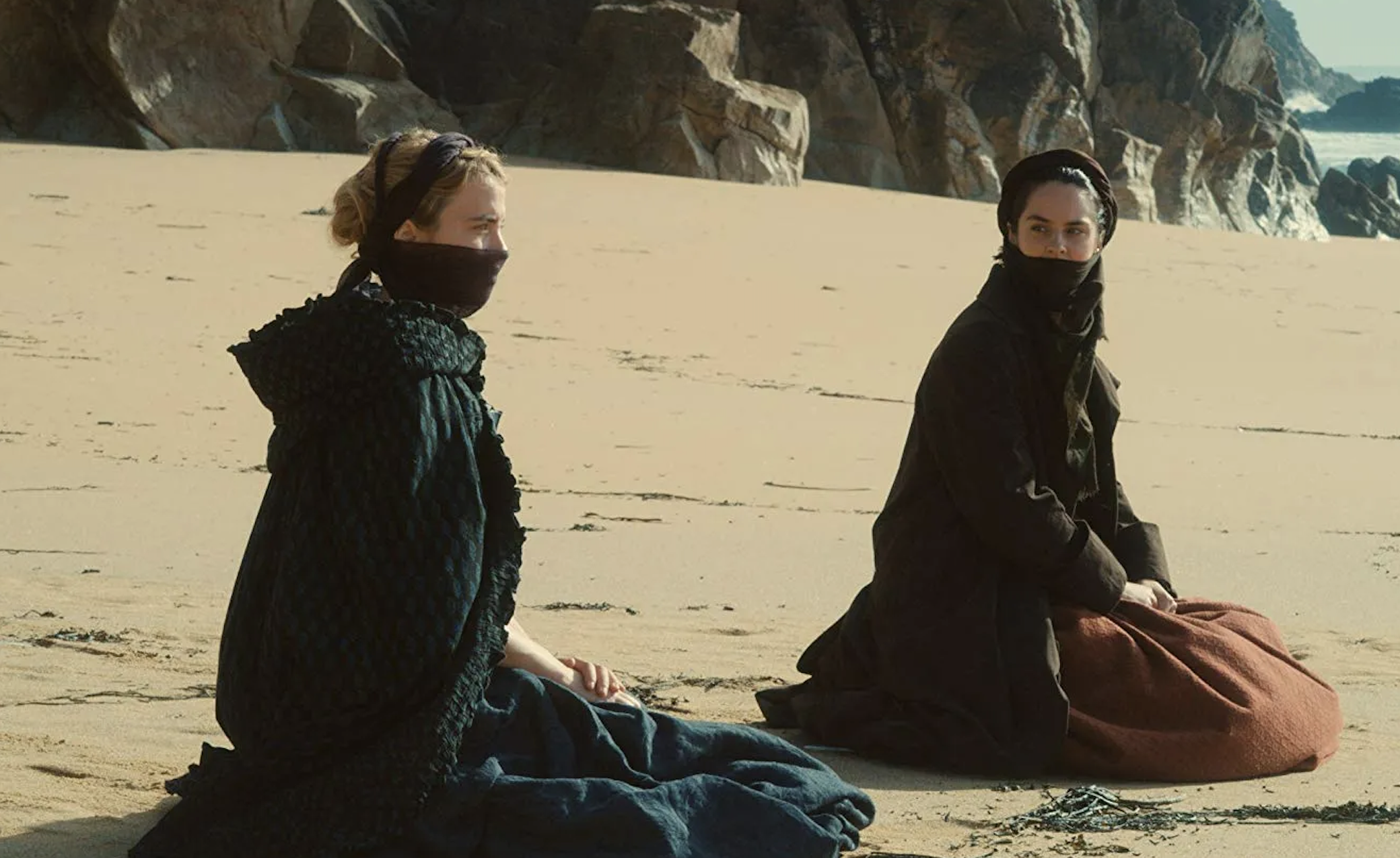 Two characters from the movie &#x27;Dune&#x27; sit on a sandy landscape, dressed in textured, dark garments
