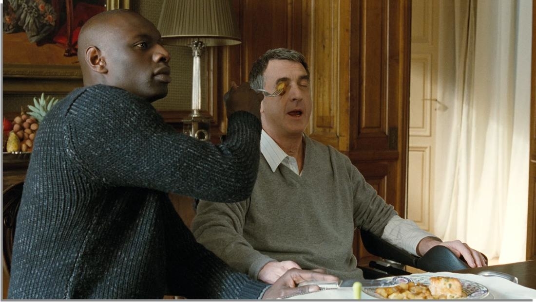 Two characters from the film &#x27;The Intouchables&#x27; sitting at a dining table, one assisting the other with eating