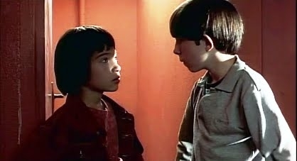 Two children having a conversation in a hallway, one with a red jacket, the other in a grey shirt