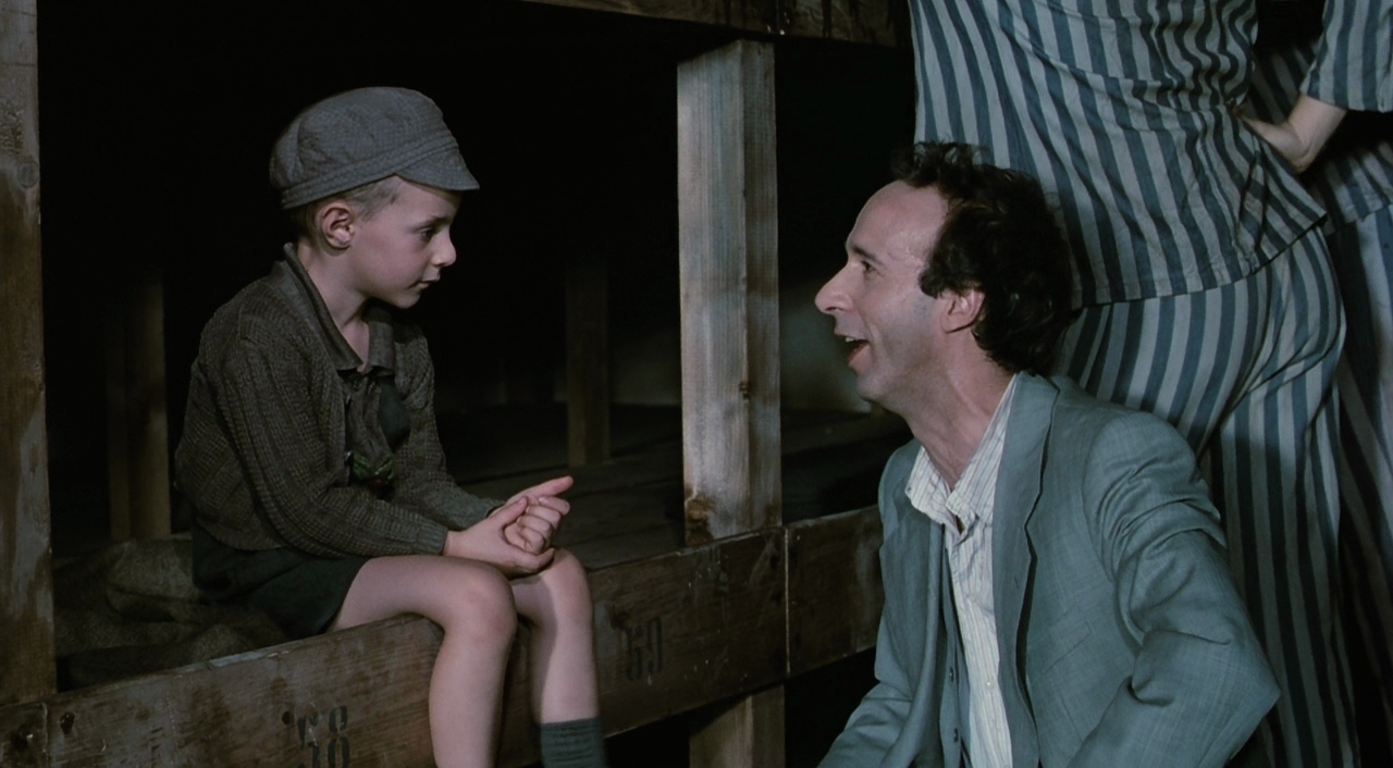 Scene from a film with a man in striped prison uniform and a boy in a cap sitting, conversing