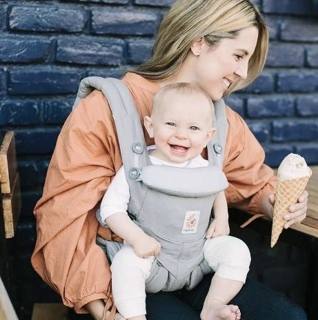 Woman seated, smiling with baby in a front carrier, both facing camera, woman holding ice cream cone