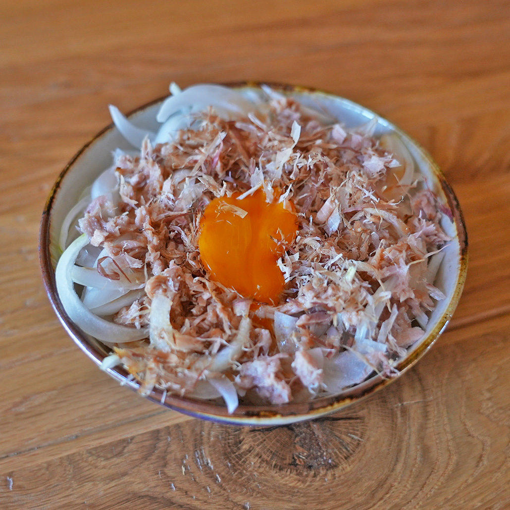 切ったタマネギの上にかつおぶしと卵黄をのせた料理が木のテーブルに置かれている。
