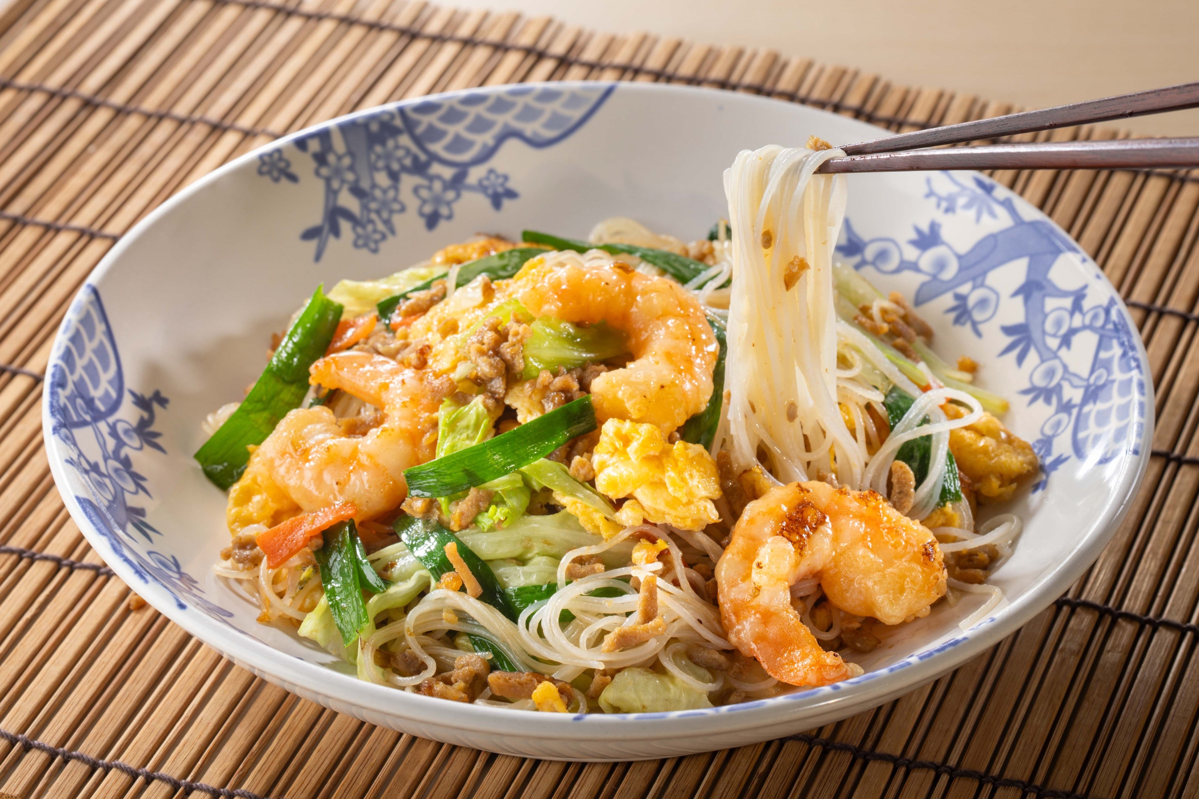 海老と野菜が入ったアジア風の麺料理を箸で持ち上げている様子。