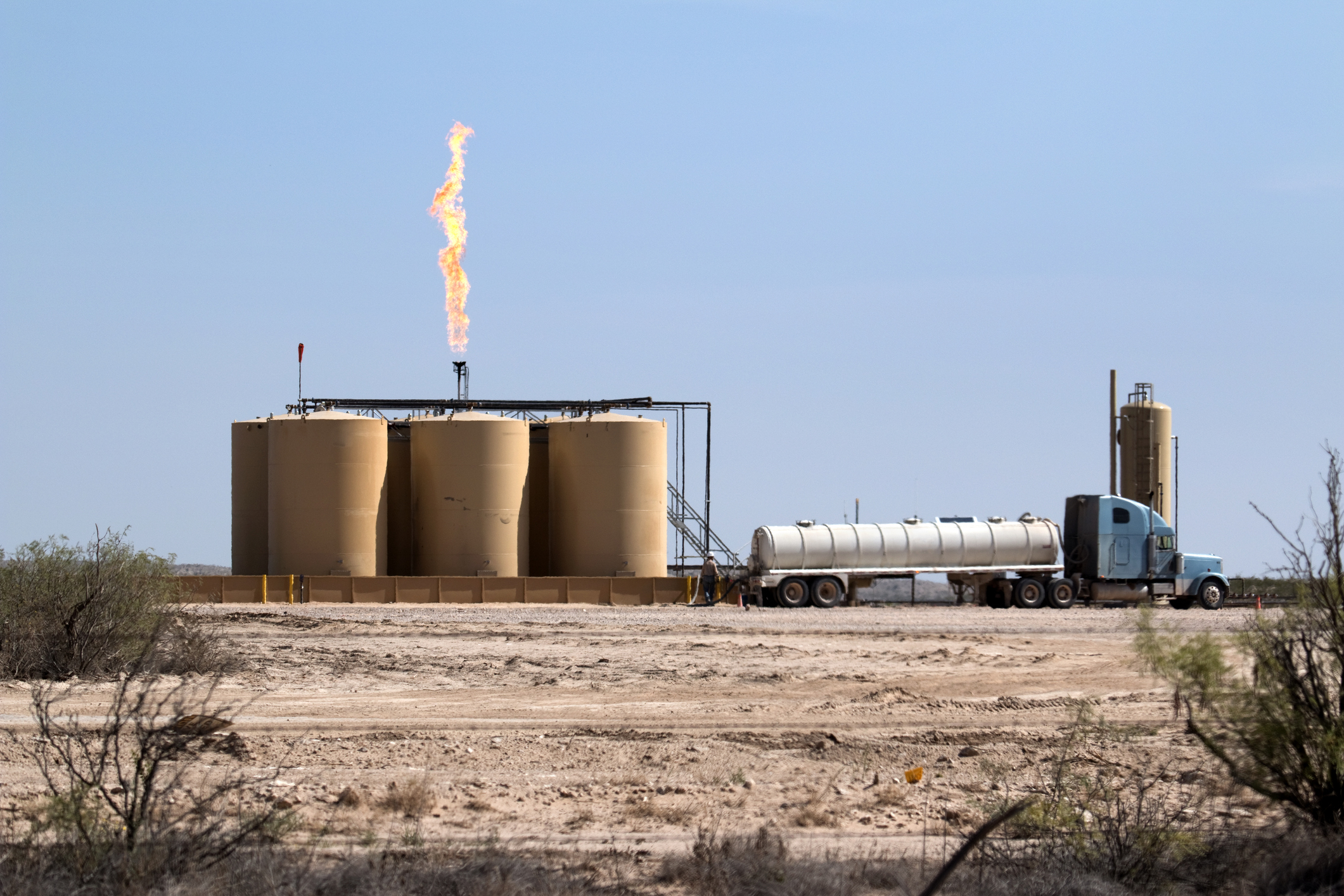 Oil refinery tanks with a flare stack burning off gas and a tanker truck parked beside in a desert area