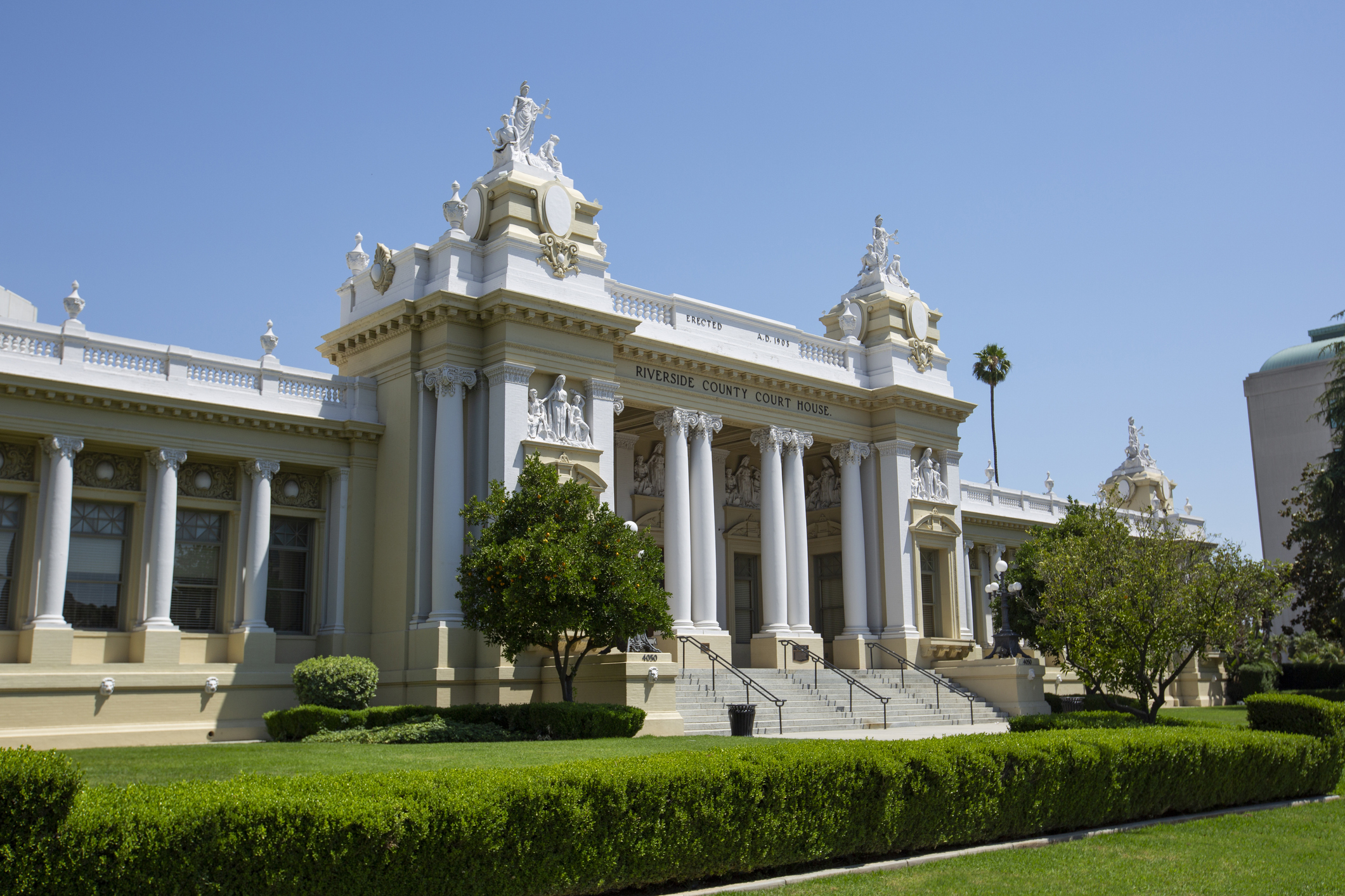 Neoclassical building with columns and sculptures atop its facade, flanked by greenery under a clear sky
