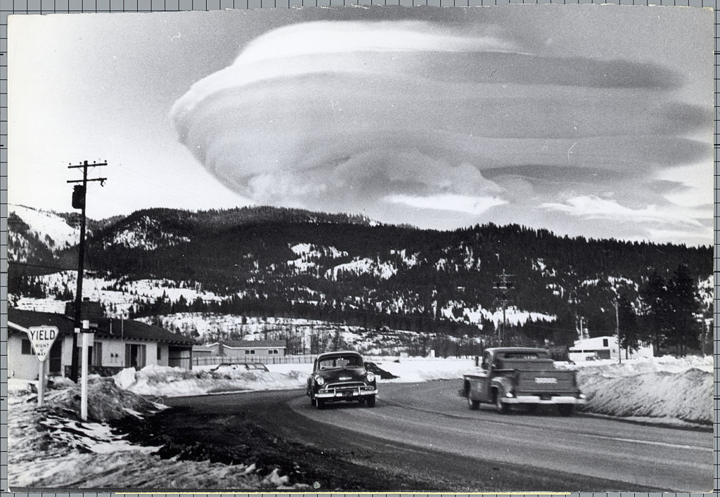Vehicles on a snowy road with a large, lenticular cloud over a mountain landscape