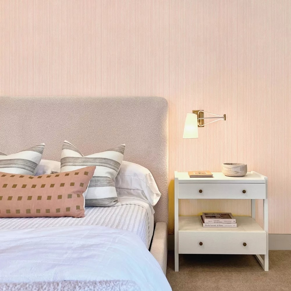 Bed with pillows against a pink wall, beside a nightstand with books and a lamp