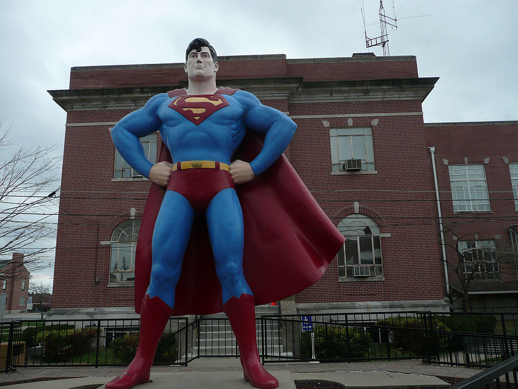 Statue of Superman, standing with hands on hips, cape flowing, against a building backdrop