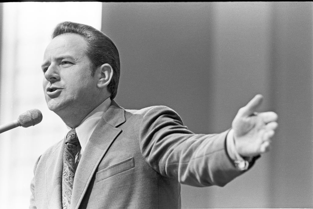 Man in a suit speaking into a microphone, gesturing with one arm outstretched