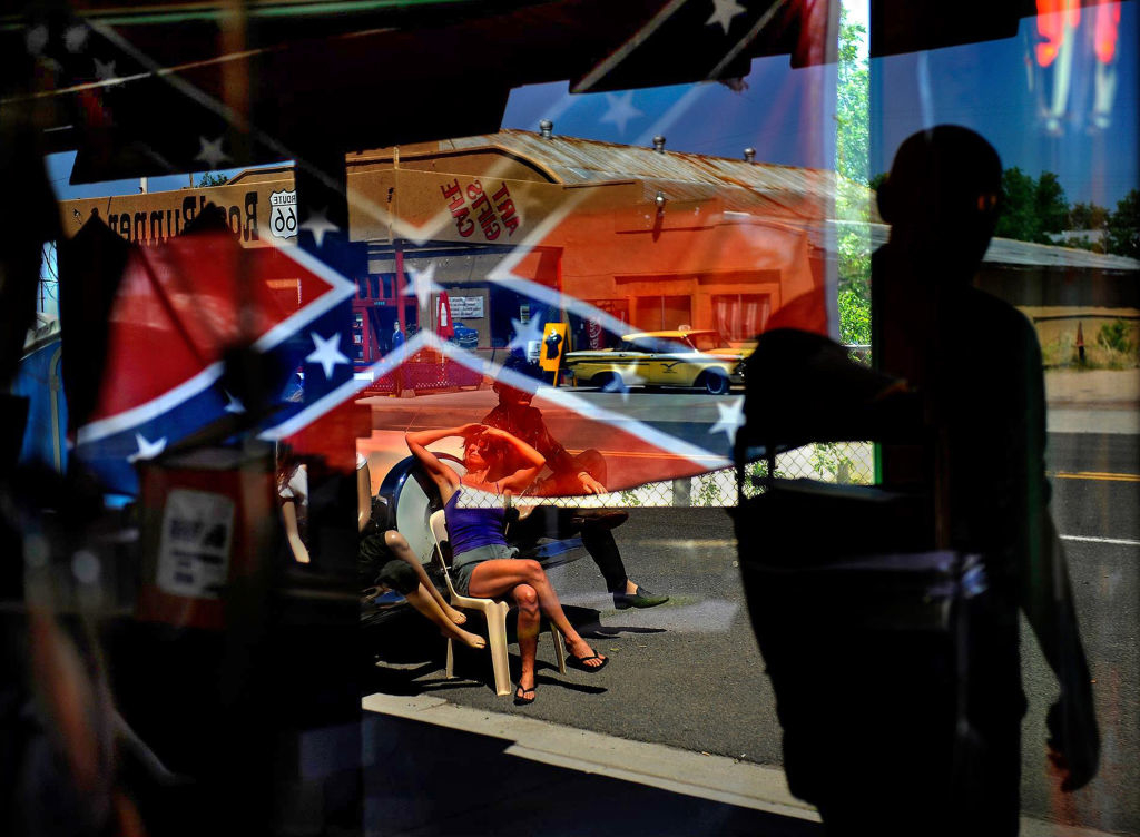 A person relaxes in a chair on a sidewalk reflected in a window adorned with Confederate flags