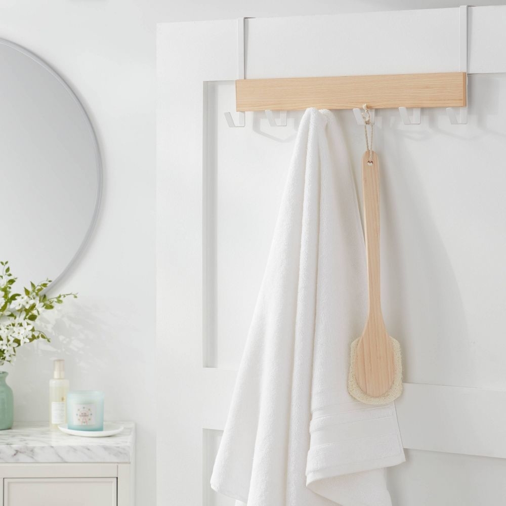 A white towel hangs on a wooden rack with a bath brush, beside a mirror and bathroom products