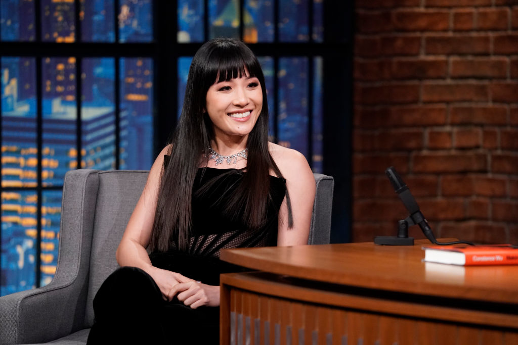 Woman in a black dress and necklace smiles seated on a talk show set