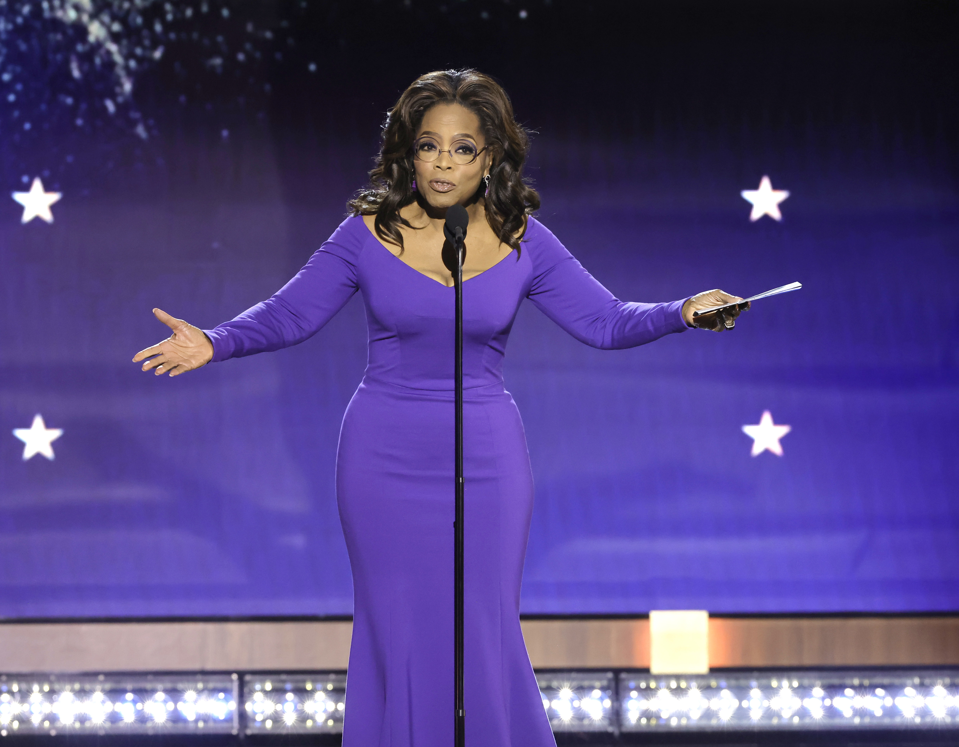 Oprah Winfrey in a long-sleeved dress, speaking at a podium onstage with stars in the background