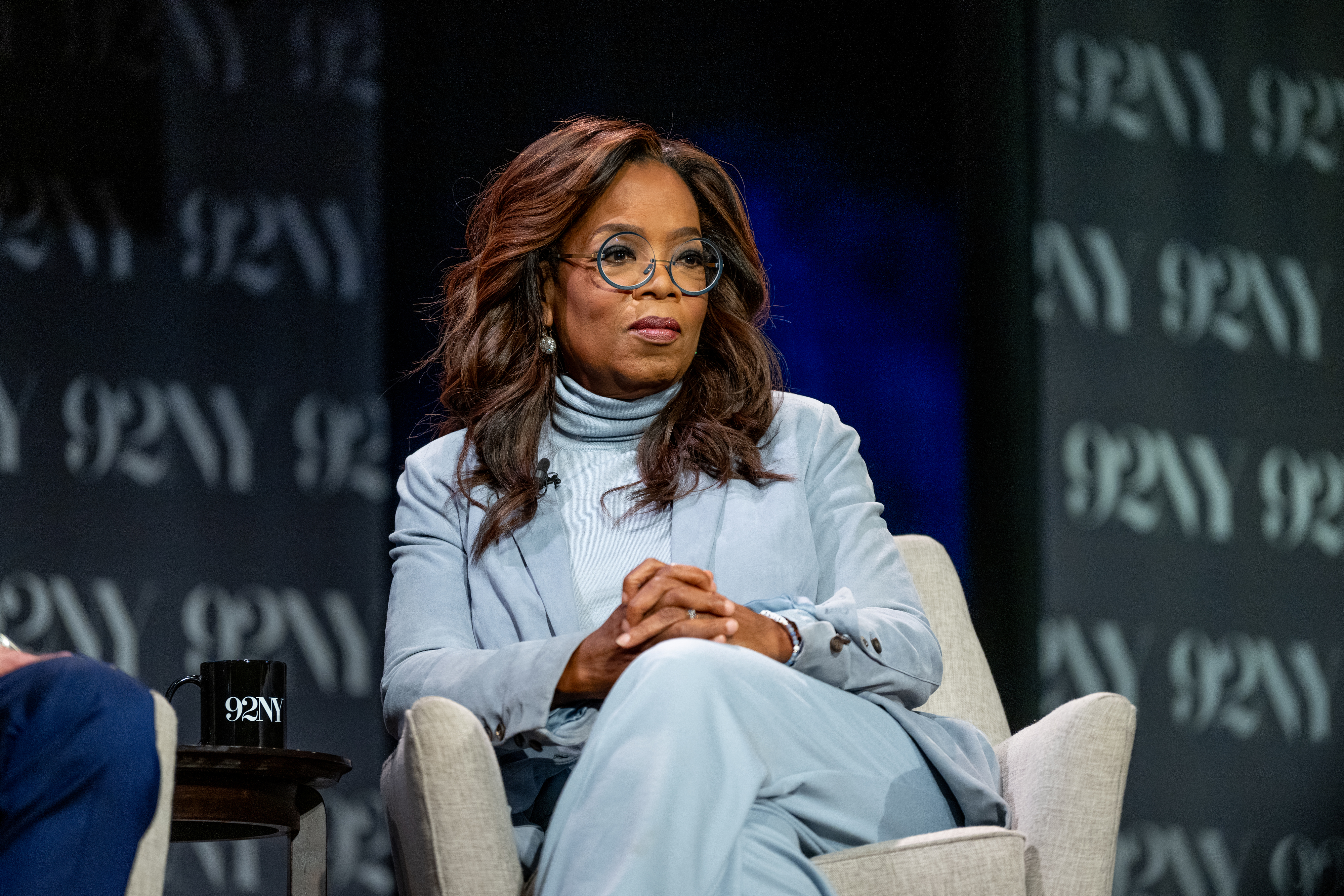 Oprah Winfrey in a panel discussion, wearing a tailored suit, seated with hands crossed
