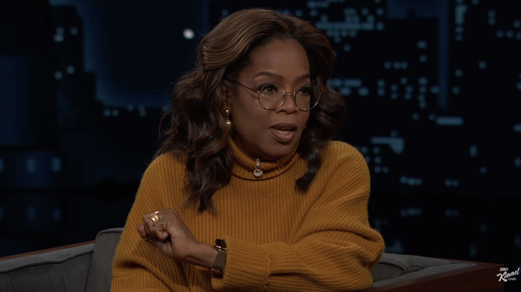 Oprah Winfrey in a sweater seated on a talk show set with a cityscape background