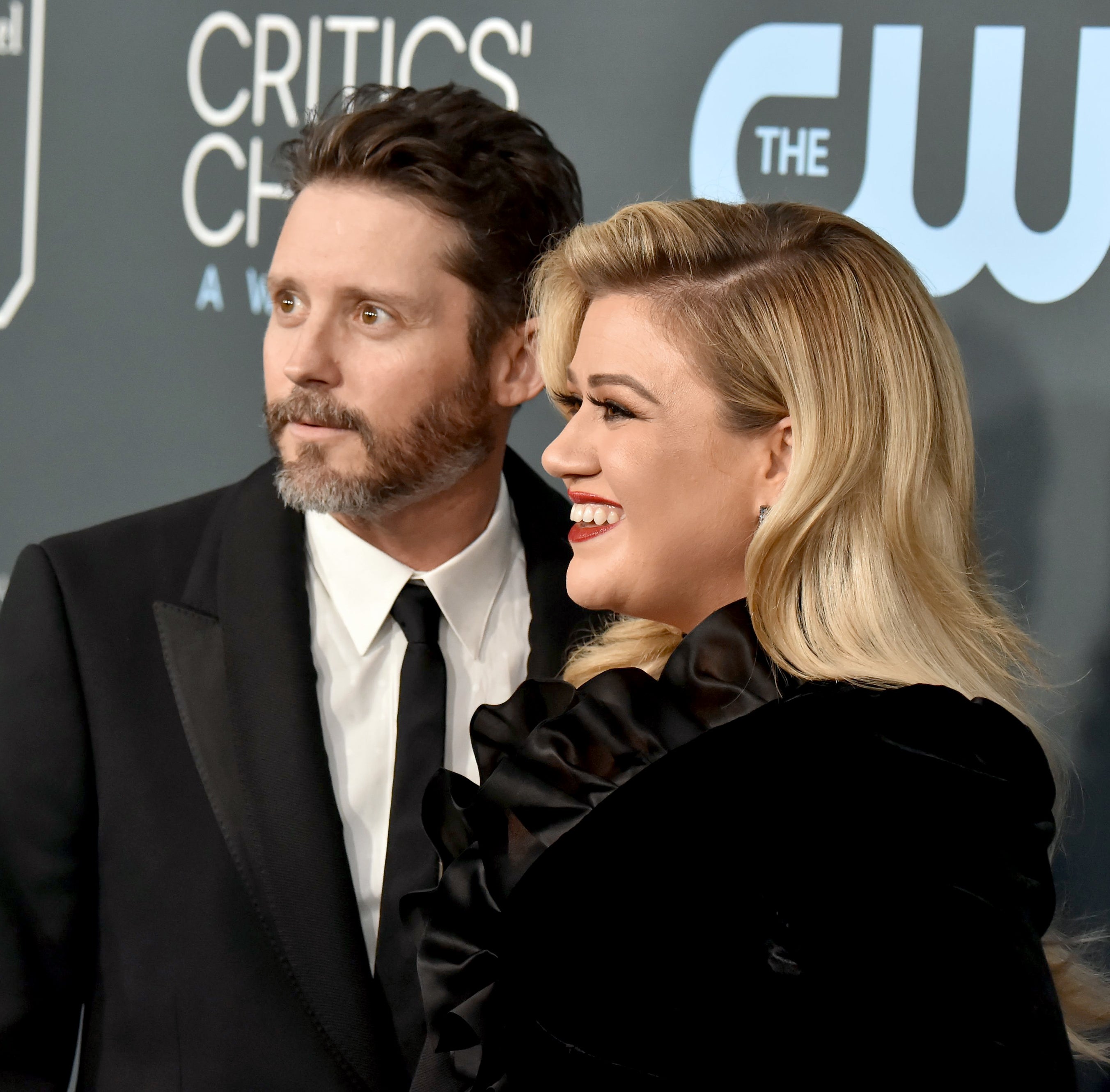 Brandon in a suit and Kelly in a dress with ruffled collar at a media event