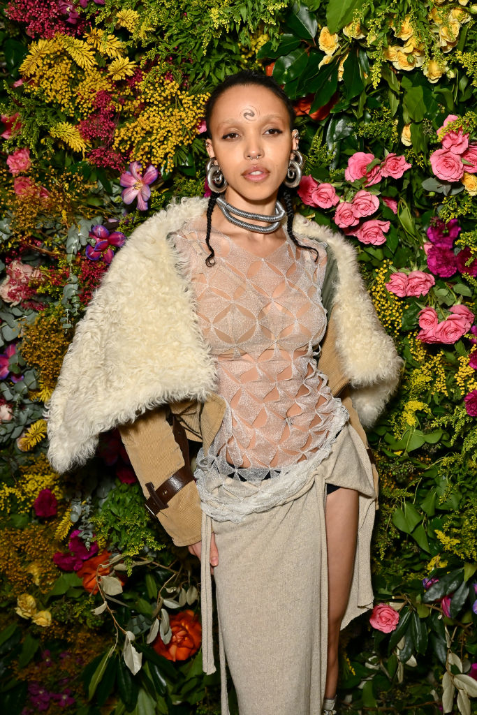 FKA Twigs posing against a floral backdrop wearing a sheer top with a geometric pattern, a cream jacket, and a draped skirt
