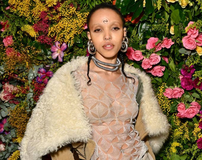 FKA Twigs posing against a floral backdrop wearing a sheer top with a geometric pattern, a cream jacket, and a draped skirt