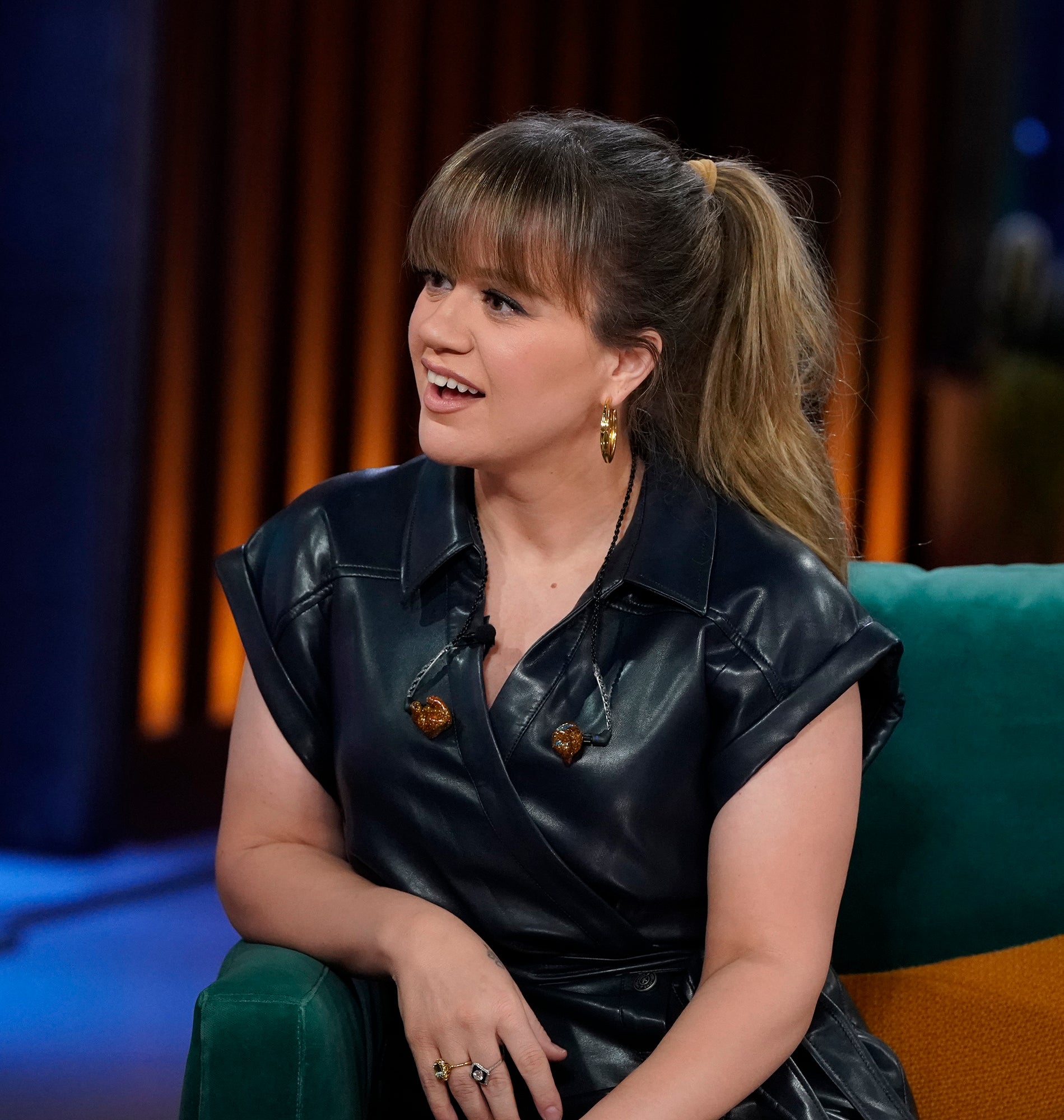 Kelly in a leather dress seated with a ponytail, on a talk show set