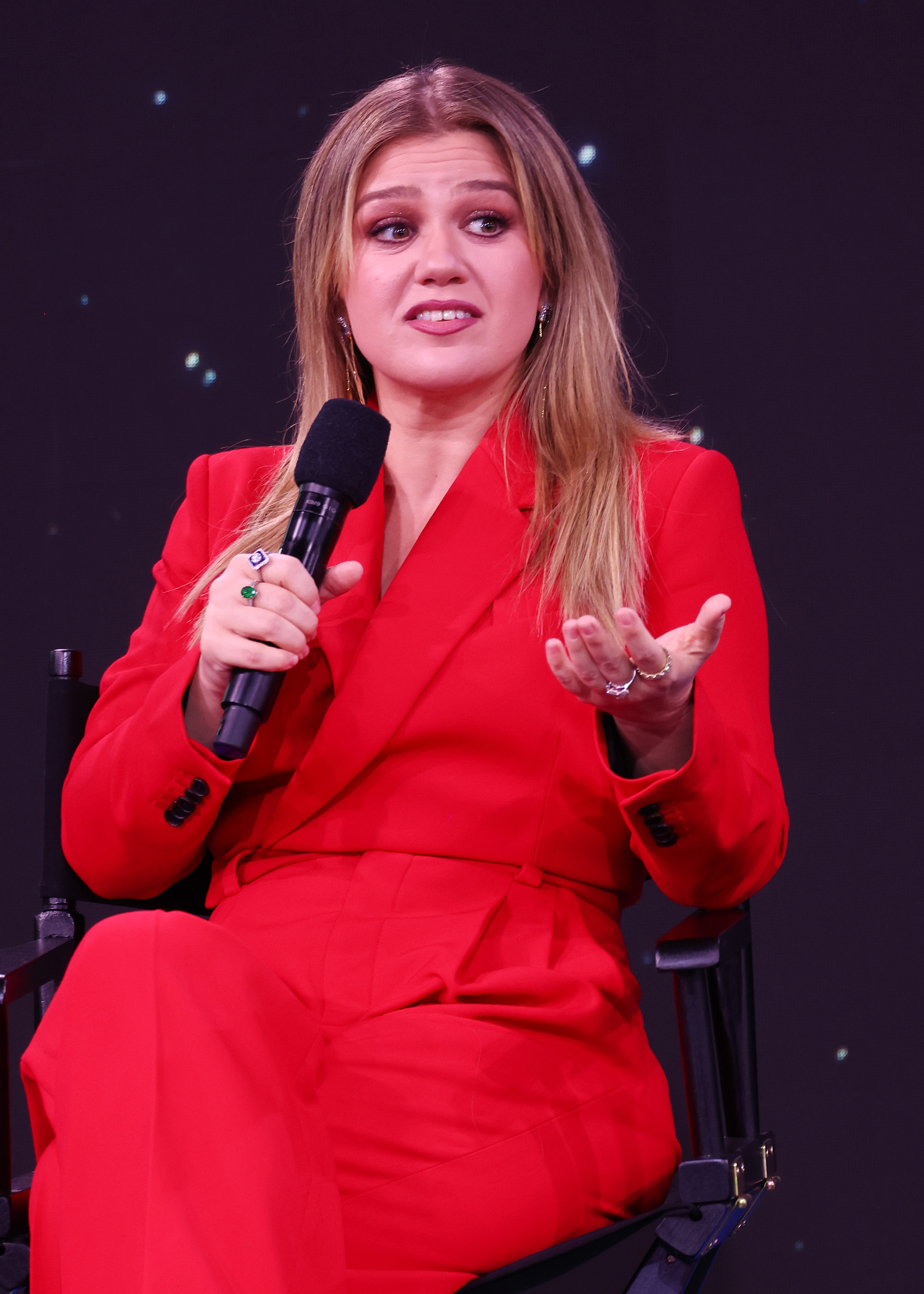Kelly in a suit sitting and speaking into a microphone