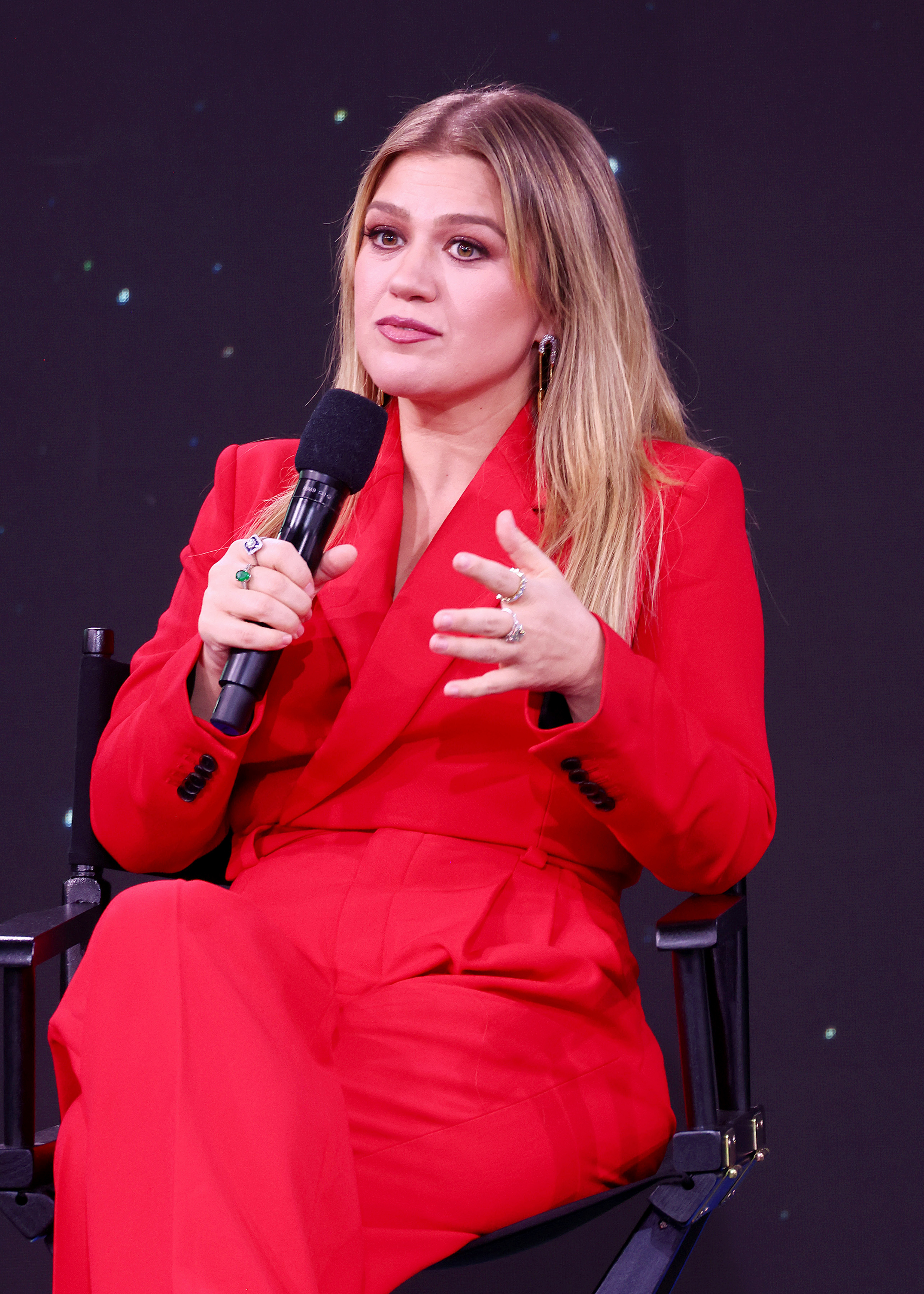 Kelly in a red suit speaking into a microphone during an event