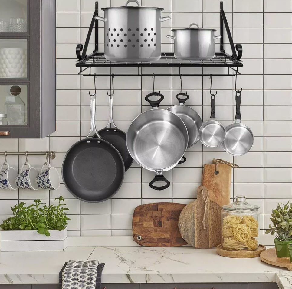 A kitchen wall with a hanging rack holding various pots, pans, and utensils, beside a shelf with plants and cutting boards